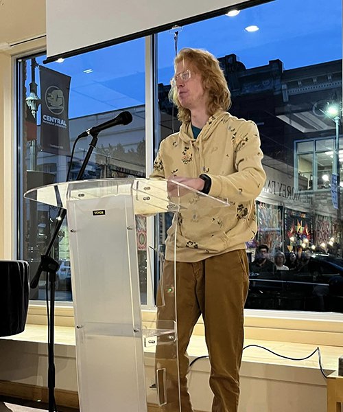 Weston Wise stands at a podium in front of glass windows to read from his poetry at Art Reach of Mid Michigan.