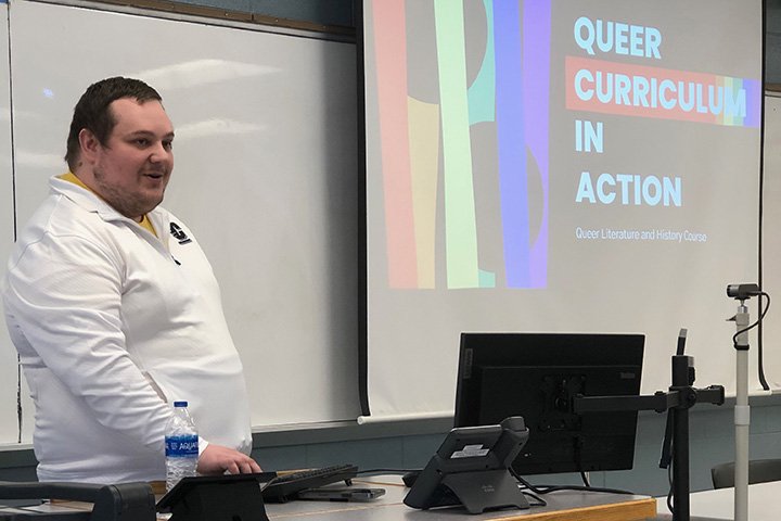 Jason Westra-Hall wearing a white Central Michigan University shirt stands between a desk and display screen in a CMU classroom.