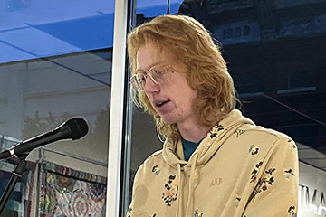 Weston Wise stands at a podium in front of glass windows to read from his poetry at Art Reach of Mid Michigan.