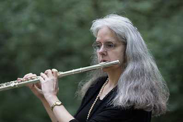 Joanna White plays a flute while standing outdoors near trees.