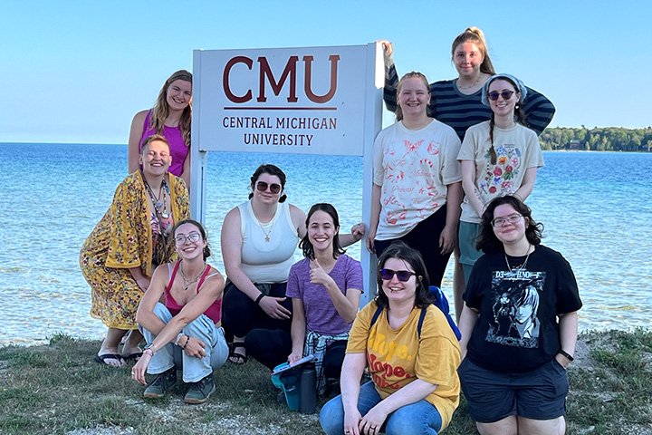 Central Michigan University students stand and sit next to a Central Michigan University sign next to Lake Michigan on Beaver Island.