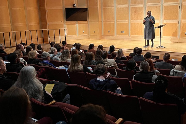 Michigan Poet Laureate Nandi Comer speaks into a microphone while standing on a stage in front of a seated audience.