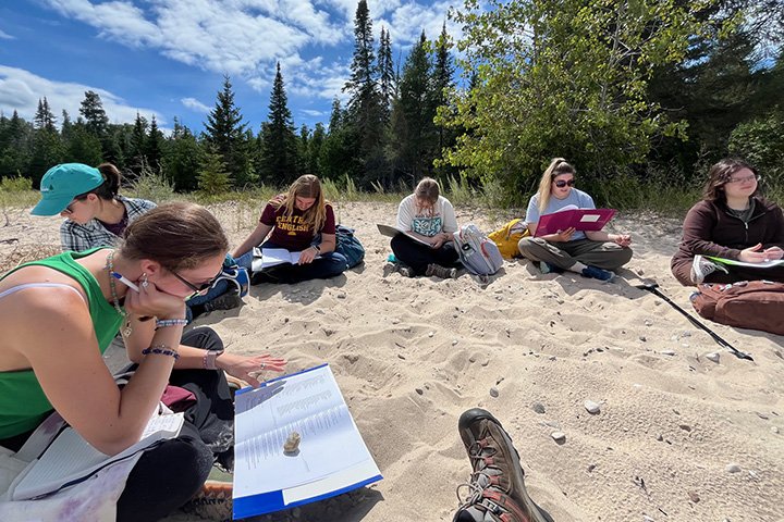 Six students read from papers in folders while seated on a sandy beach.