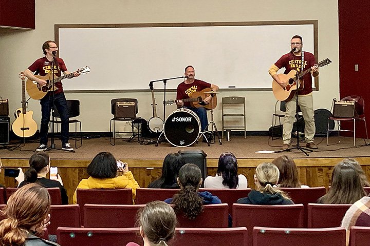 English Department faculty members (from left to right) Jeffrey Bean, Darrin Doyle, and Robert Fanning play guitars and drums on stage while student watch from chairs in a lecture hall.