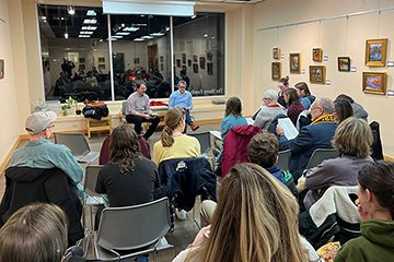 English faculty members Nate Smith and Mark Freed seated in chairs next to large windows facing people sitting in several rows of chairs.