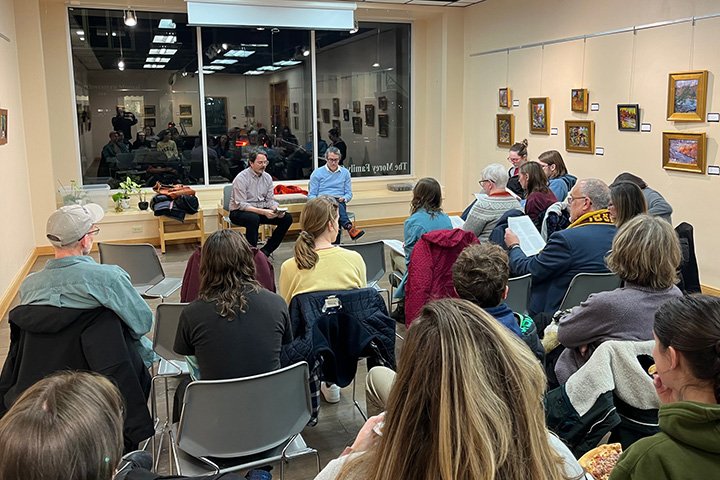 English faculty members Nate Smith and Mark Freed seated in chairs next to large windows facing people sitting in several rows of chairs.
