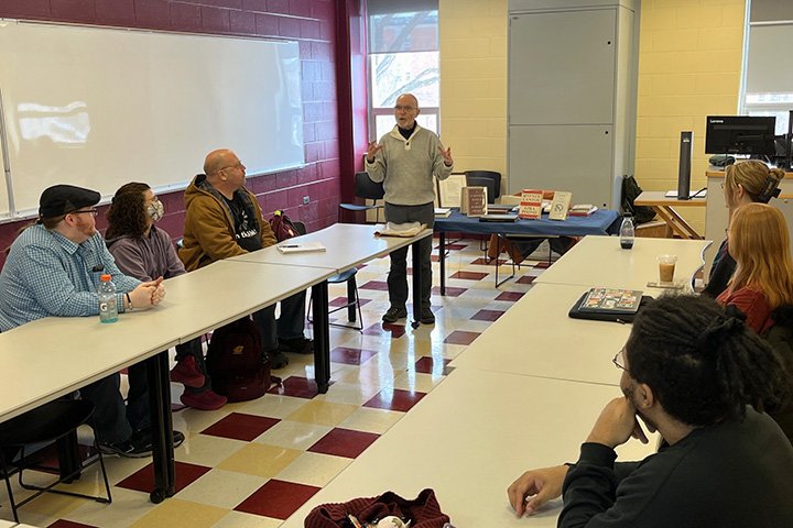 Jett Whitehead stands near table filled with rare books while students are seated at long tables lining two sides of the classroom.