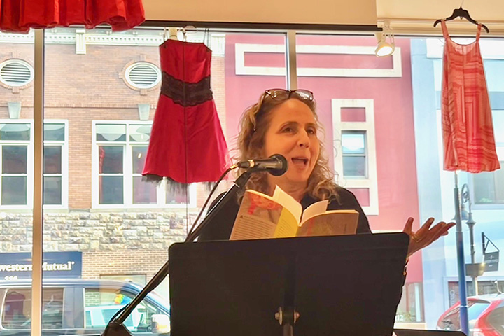 Poet Alise Alousi holds a book and reads poetry while standing in front of a large glass window at Art Reach of Mid Michigan