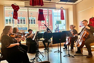 A string sextet featuring Central Michigan University School of Music students plays while seated in chairs in front of a large glass window at Art Reach of Mid Michigan