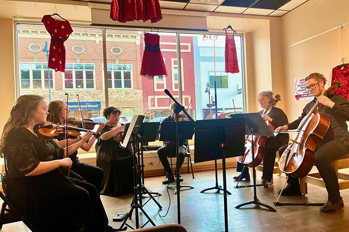 A string sextet featuring Central Michigan University School of Music students plays while seated in chairs in front of a large glass window at Art Reach of Mid Michigan