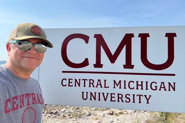 English Professor Robert Fanning wearing a hat and sunglasses stands next to a Central Michigan University sign on Beaver Island.