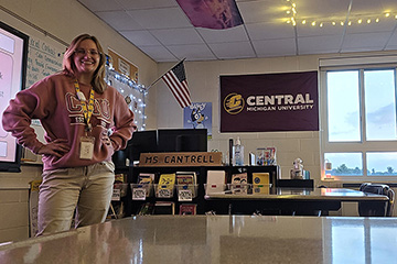 Desiree Cantrell stands in her classroom while wearing a CMU sweatshirt and with an American flag and CMU flag on the walls behind her desk.