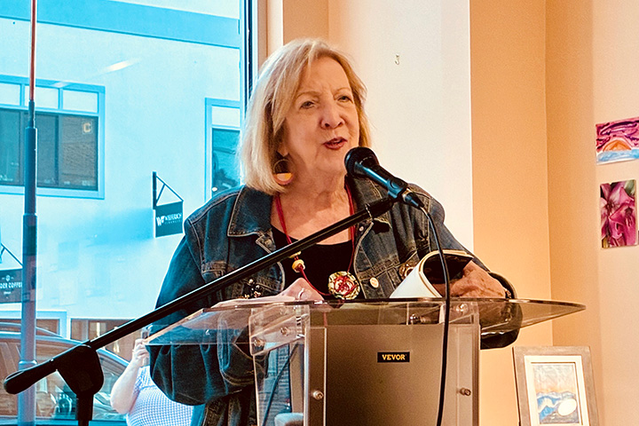 Patricia Clark speaks into a microphone while standing at a podium and flipping through pages of her book.