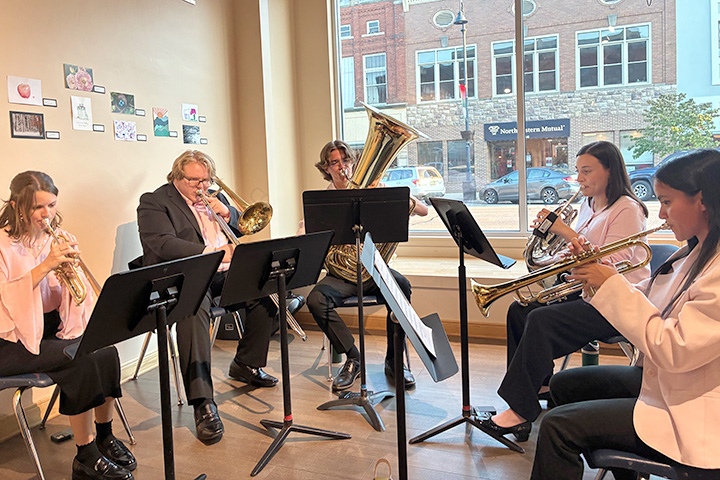 Members of the brass quintet Sassabrass play instruments while seated in chairs in a half-circle facing music stands.