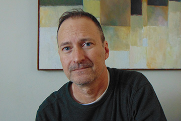 Closeup image of Darrin Doyle wearing a dark shirt and seated in front of a decorative painting