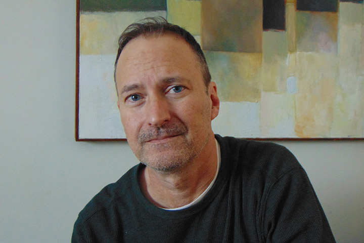 Closeup image of Darrin Doyle wearing a dark shirt and seated in front of a decorative painting