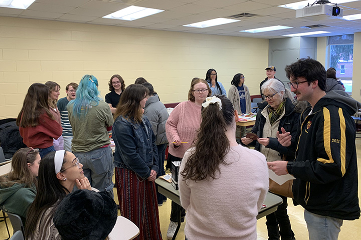 Groups of students stand around desks facing each other in a classroom to discuss items placed on the desks.