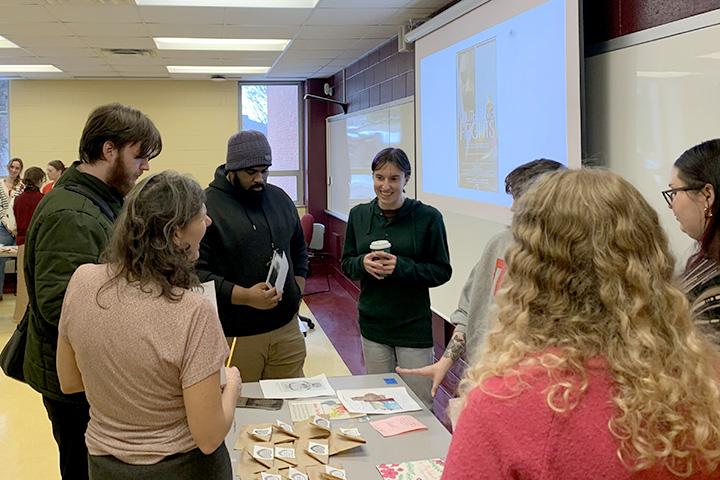 Groups of students stand around desks facing each other in a classroom to discuss items placed on the desks.