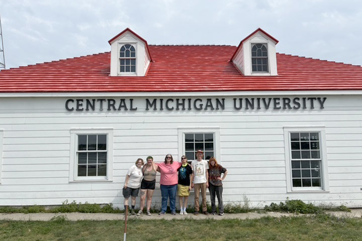Students stand in front of the Central Michigan University boathouse on Beaver Island, a large building with white siding and a red roof.