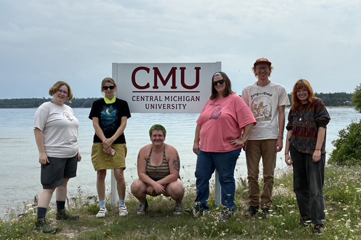 Students stand in front of a Central Michigan University sign with a lake behind them.