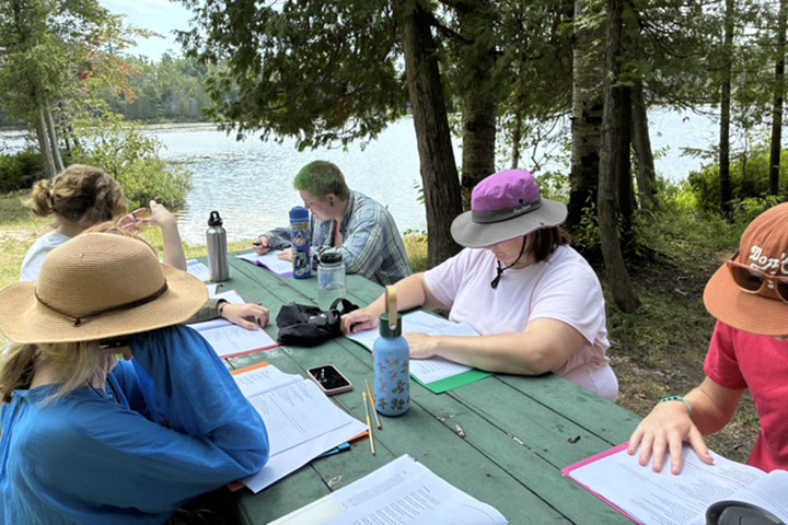 Students sit on a picnic table near a lake facing each other with books open in front of them.