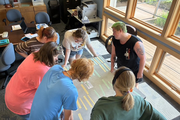Students collaborate on creating a poem with strips of paper with words laid out on a table next to a wall of glass windows.