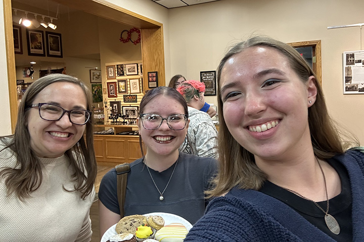 English faculty member Holly Lewis stands near two students with one holding a plate of cookies and desserts.