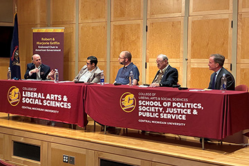 Griffin Forum panelists are seated at two tables with maroon CMU table cloths on a wooden stage.