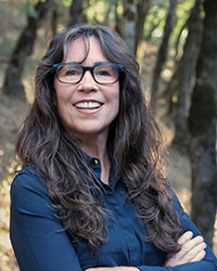Image of Kori Schake wearing a blue shirt and glasses with her arms crossed