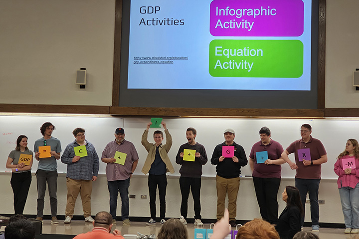 10 individuals stand in a row across a stage while holding a square paper with letters or symbols written on them.