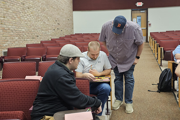 One individual stands and looks over the shoulder of another individual seated at a desk while reviewing pieces of paper as a third seated individual observes.