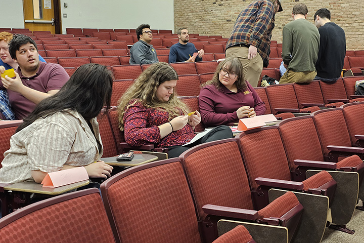 Multiple individuals are seated in rows of red chairs in an auditorium while holding small papers.