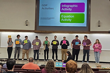 10 individuals stand in a row across a stage while holding a square paper with letters or symbols written on them.