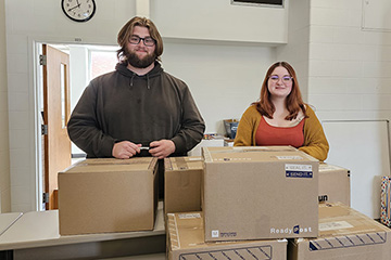 Nicholas Nance (left) and Lauren Cole stand behind stacks of brown shipping boxes filled with donated school supplies.