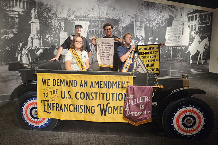 Students pose behind a prop car with bold yellow signs and banners from the 19th Amendment exhibit