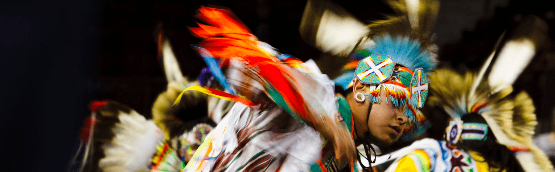 A Native American dancer in colorful regalia performs at a powwow. The motion blur captures the energy of the dance, highlighting feathered headdresses, beadwork, and bright fabric in red, blue, yellow, and green.