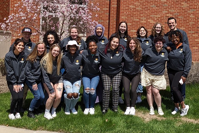 Intergroup Dialogue Facilitators and faculty members stand in two rows in front of flowering tree next to Anspach Hall.