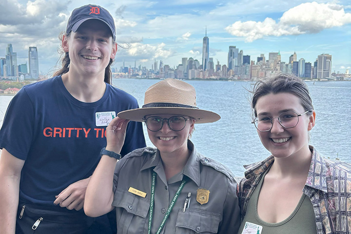 CMU alum Fern Borus wearing National Park Service uniform and hat stands between CMU alumni Noah Walther and Trinity Sapp with water and the New York City skyline behind them.