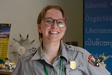 Image of Fern Borus wearing a National Park Service uniform while standing at the information desk for the Statue of Liberty National Monument.
