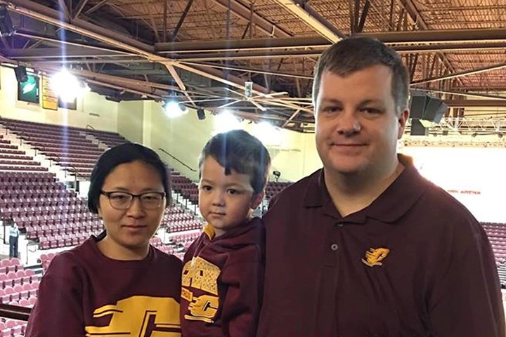 Josh Adams wearing a maroon CMU shirt stands with his wife and son inside McGuirk Arena.