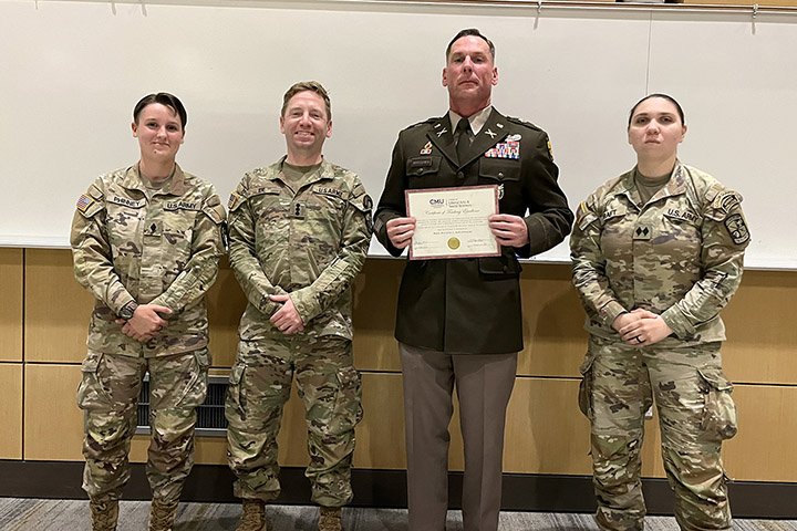 Major Nicholas J. Andrychowski holds an award while standing with three cadets wearing military uniforms.