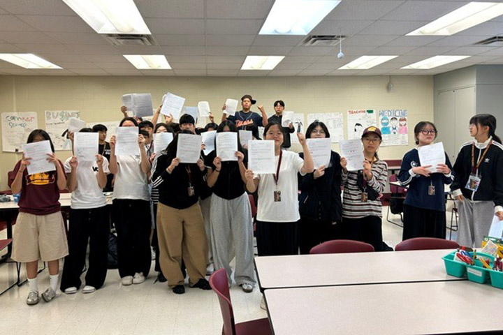 Students hold pieces of paper while standing in rows inside a classroom with desks and chairs