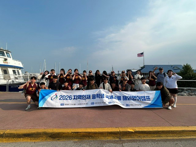 Students stand in kneel in two rows holding a banner near water and a Mackinaw Island ferry.