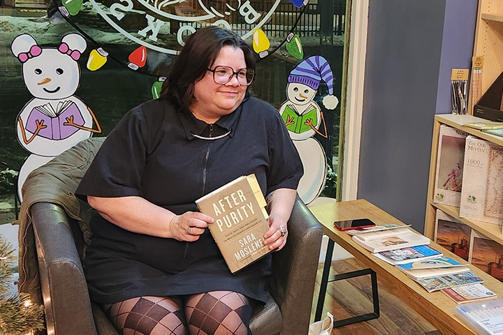 Sara Moslener holds her new book After Purity while seated in a chair next to a wooden table lined with books.