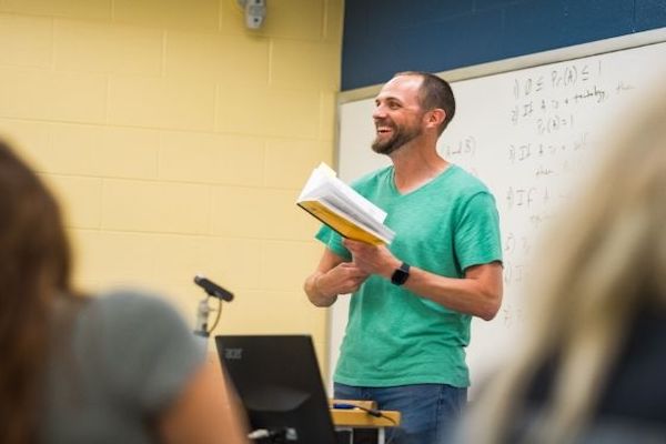 A professor holding a book at the front of the classroom and smiling at the class.