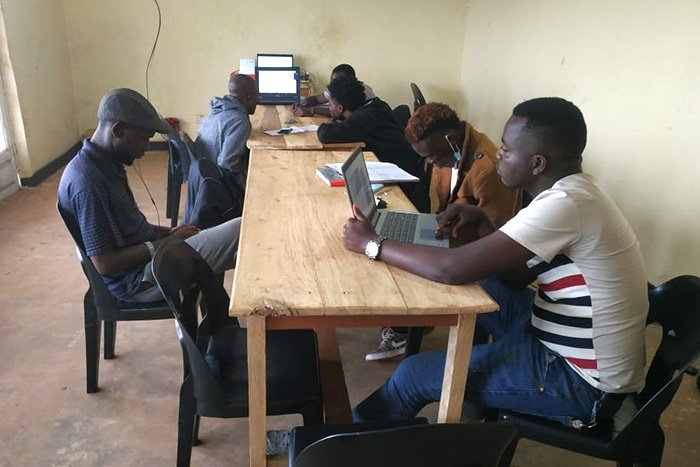 Six Global Classroom students with laptops and books are seated on both sides of two long wooden tables.