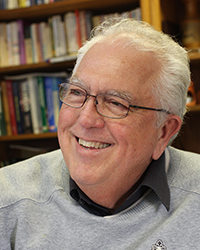 Closeup image of Del Ringquist wearing a grey sweater over a black button-up shirt while seated in front of bookshelves.
