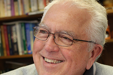 Closeup image of Del Ringquist wearing a grey sweater over a black button-up shirt while seated in front of bookshelves.