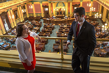 Two individuals wearing business attire talk while standing on a balcony inside the Michigan State Capitol.