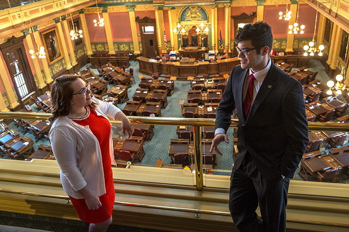 Two individuals wearing business attire talk while standing on a balcony inside the Michigan State Capitol.